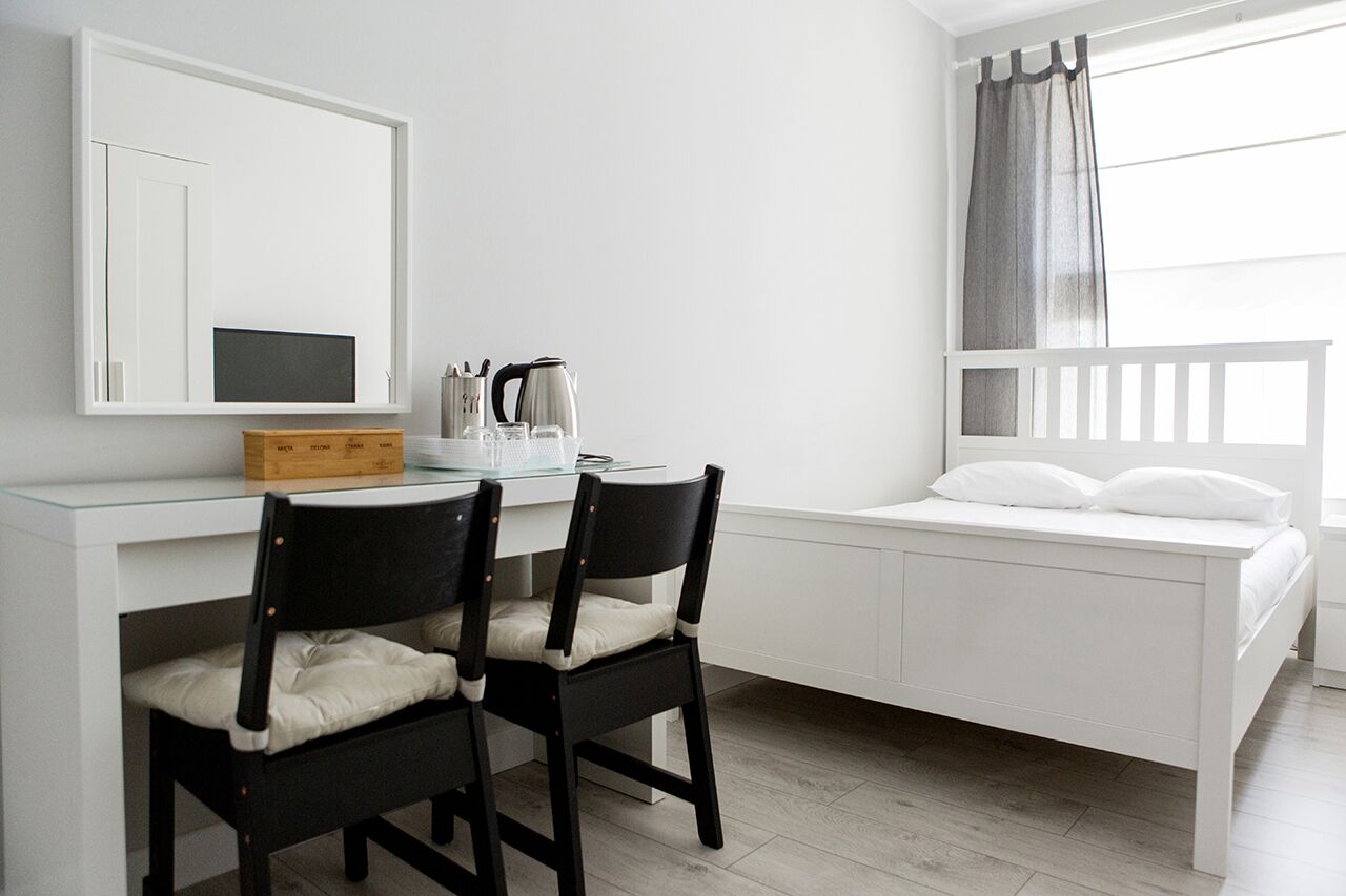 A small, modern room with a white desk, two chairs, a bed with white bedding, a large mirror, and a window with curtains.