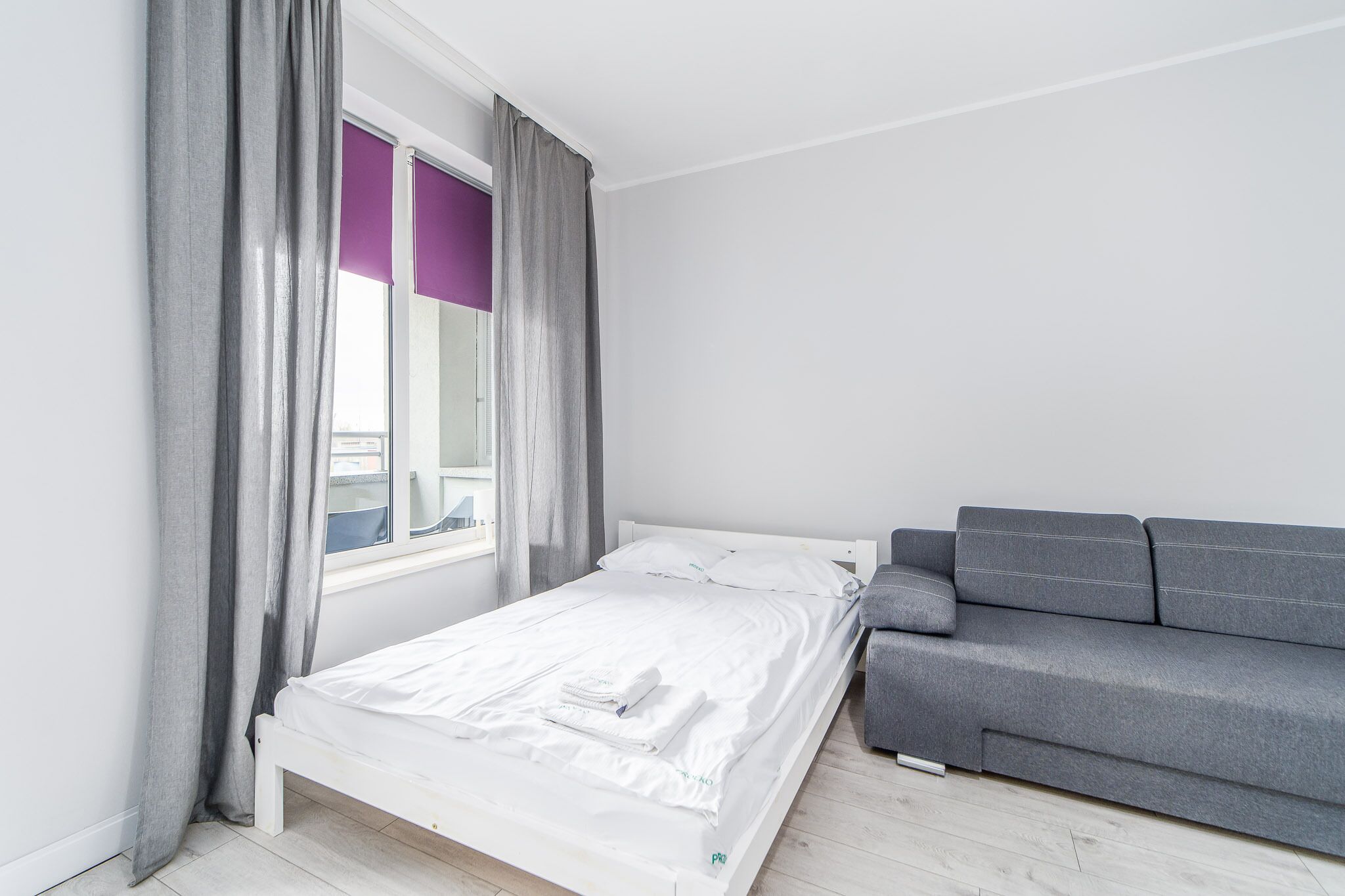 Un dormitorio con cama, cortinas grises, un sofá gris y una ventana con persianas moradas.