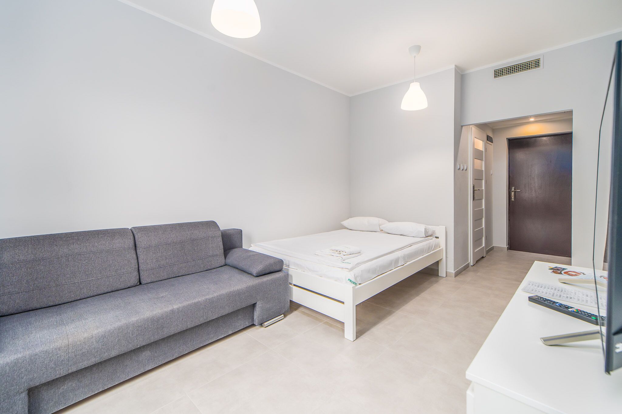 Una habitación moderna y minimalista con un sofá gris, una cama individual, un escritorio blanco con una computadora y una puerta que da a un armario.