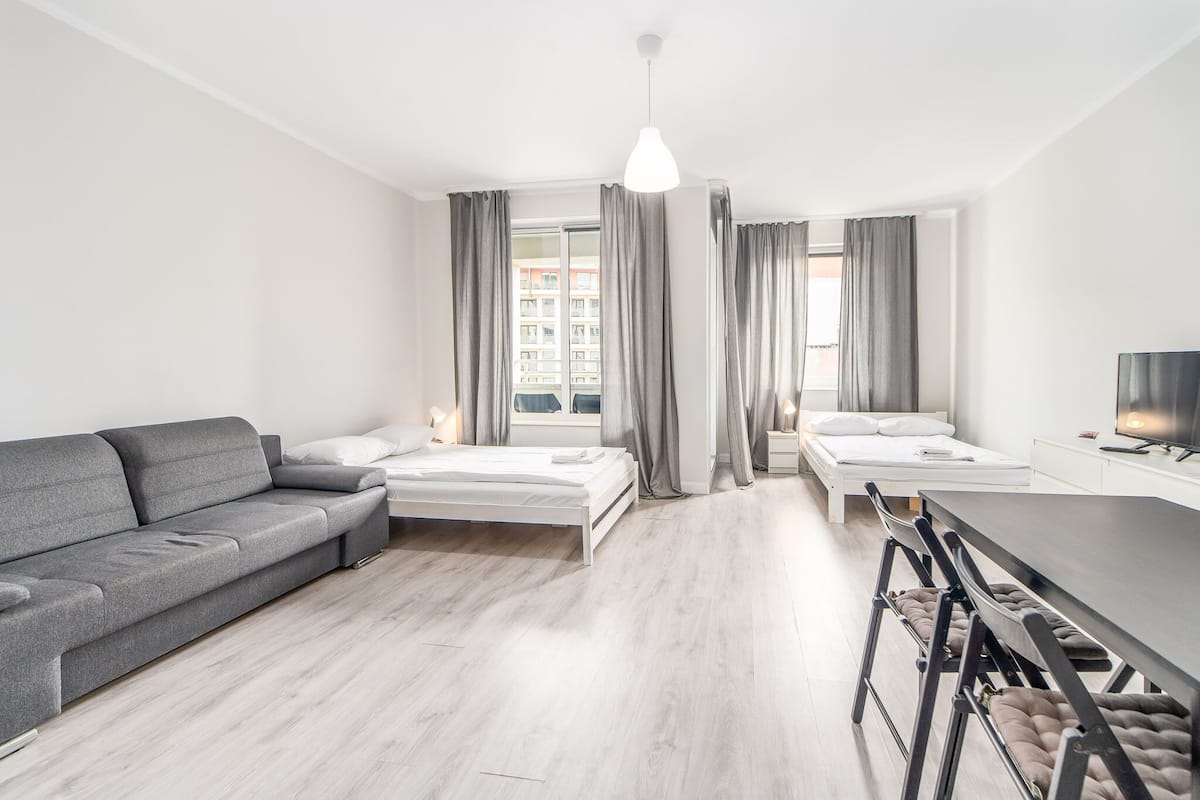 Ein moderner, gut beleuchteter Raum mit grauem Sofa, einem Bett, einem Esstisch mit Stühlen und einem Fernseher.