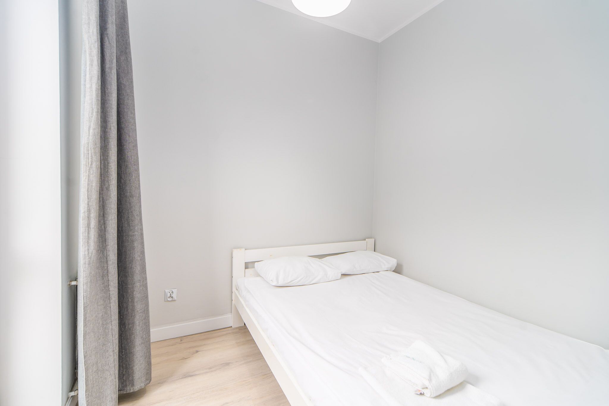 Una habitación pequeña con cama individual, cabecero blanco, suelo de madera y cortina gris entreabierta.