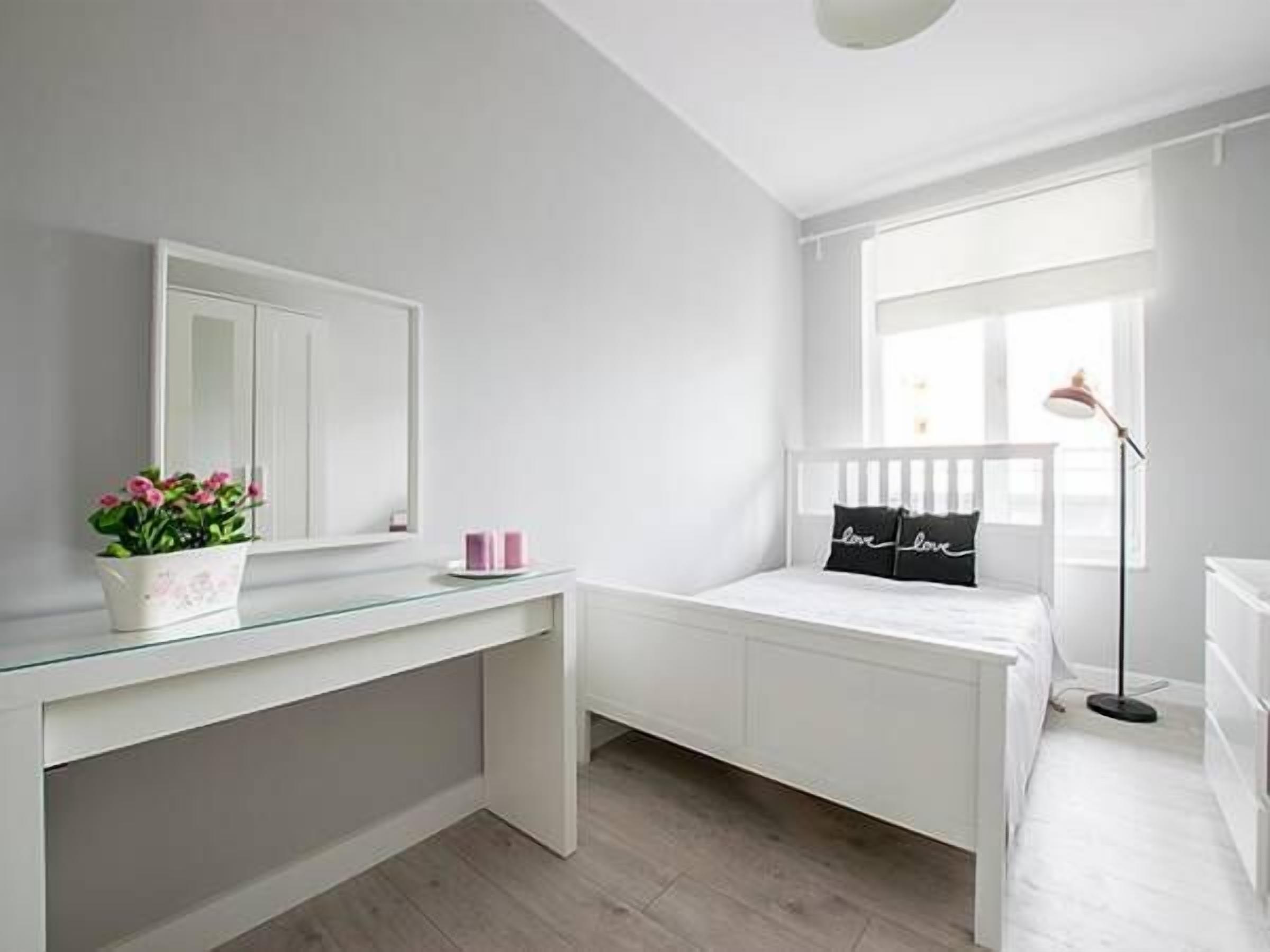 Un dormitorio con una cama blanca, una mesita de noche, un espejo, una ventana y una lámpara de pie.