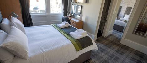 Un lit bien fait, avec du linge de lit blanc et un plaid vert, un téléviseur à écran plat fixé au mur et un tapis à carreaux.