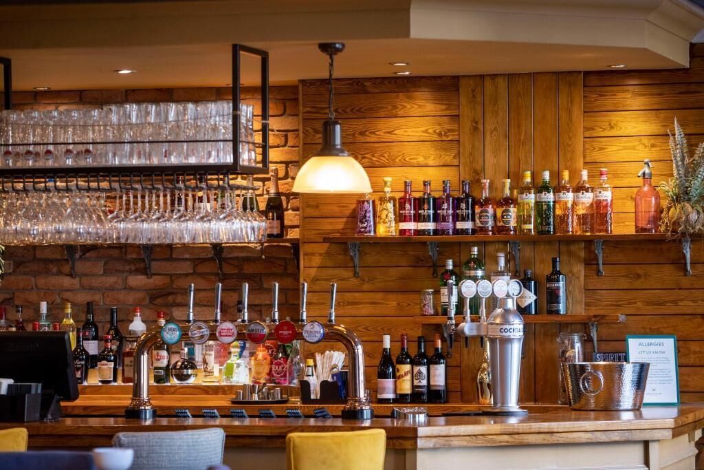 Zona de bar con pared de madera, estantes con botellas y barra con grifos y vasos.