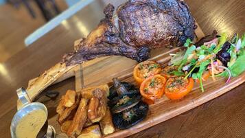 A wooden serving board with a roasted lamb shank, roasted potatoes, grilled tomatoes, and a salad.