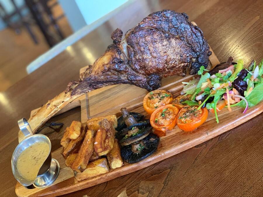 A wooden serving board with a roasted lamb shank, roasted potatoes, grilled tomatoes, and a salad.