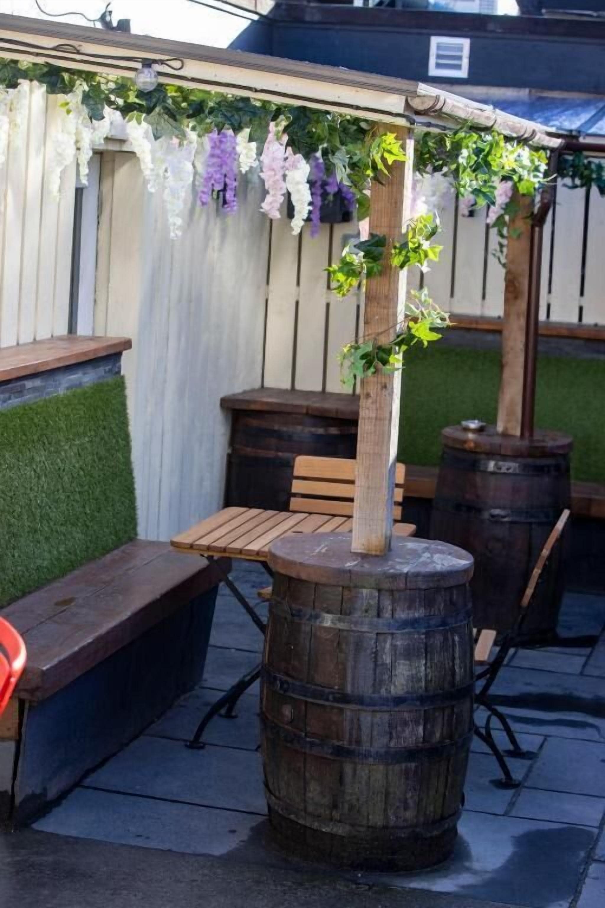Un barril de madera usado como mesa, con un banco y sillas alrededor, una pérgola con plantas colgantes y una cerca de madera al fondo.