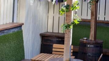 Un barril de madera usado como mesa, con un banco y sillas alrededor, una pérgola con plantas colgantes y una cerca de madera al fondo.