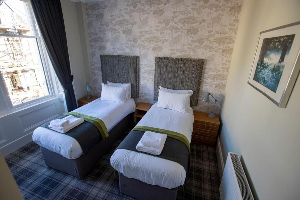 A hotel room with two beds, a window with curtains, a radiator, and a framed picture on the wall.