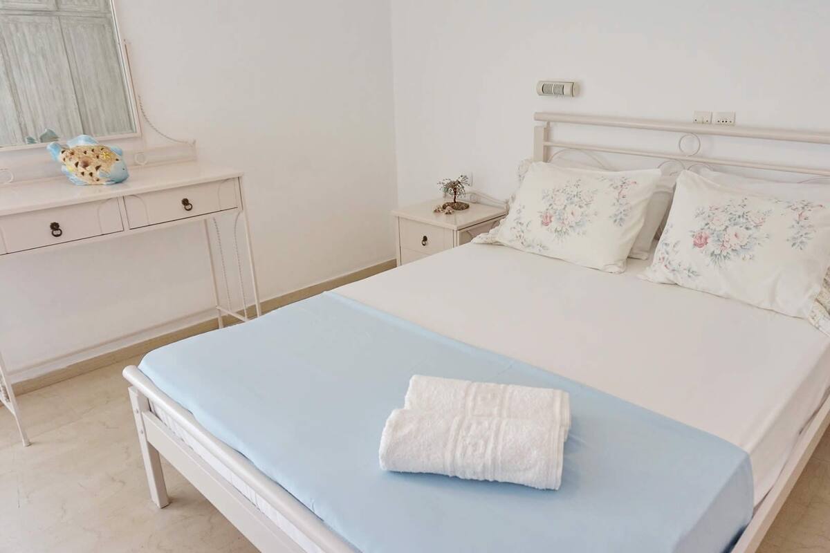 A neatly made bed with a blue bedspread and white pillows, a white dresser with drawers, and a bedside table with a lamp and decorative items.