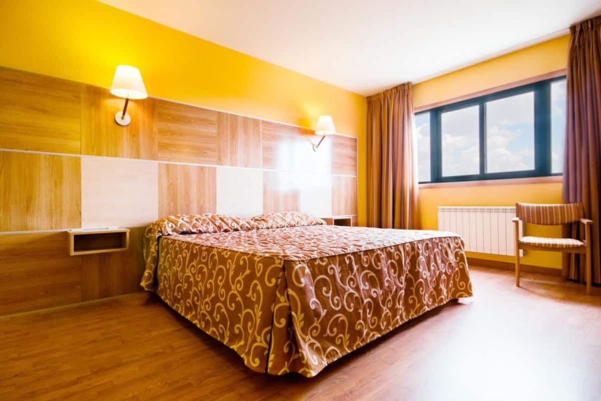 Habitación de hotel con cama, dos lámparas de pared, una ventana con cortinas y un radiador.