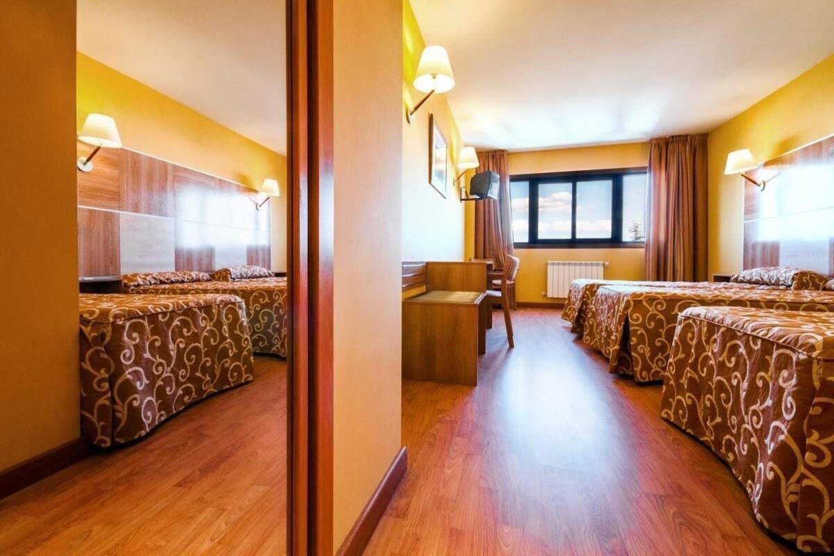 Habitación de hotel con piso de madera, dos camas con ropa de cama estampada, una mesita y una ventana con cortinas.