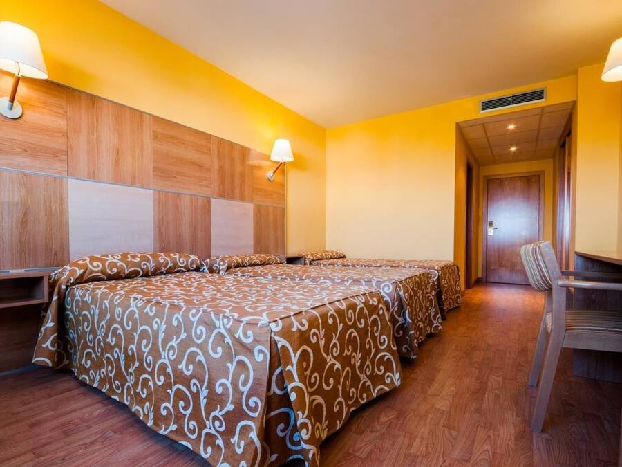 A hotel room with a wooden headboard, a bed with a patterned bedspread, a desk with a chair, and a lamp on the wall.