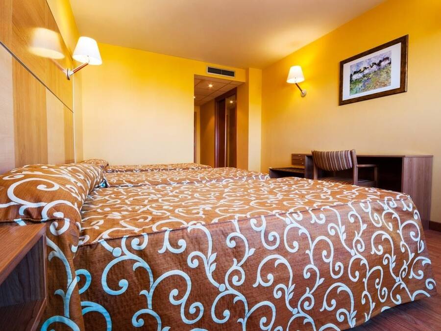 Une chambre d’hôtel avec un grand lit, un bureau et une chaise. Il y a un tableau au mur et une lampe sur le bureau.