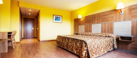 Habitación de hotel con piso de madera, cama con colcha estampada, escritorio con silla y lámpara de pared.