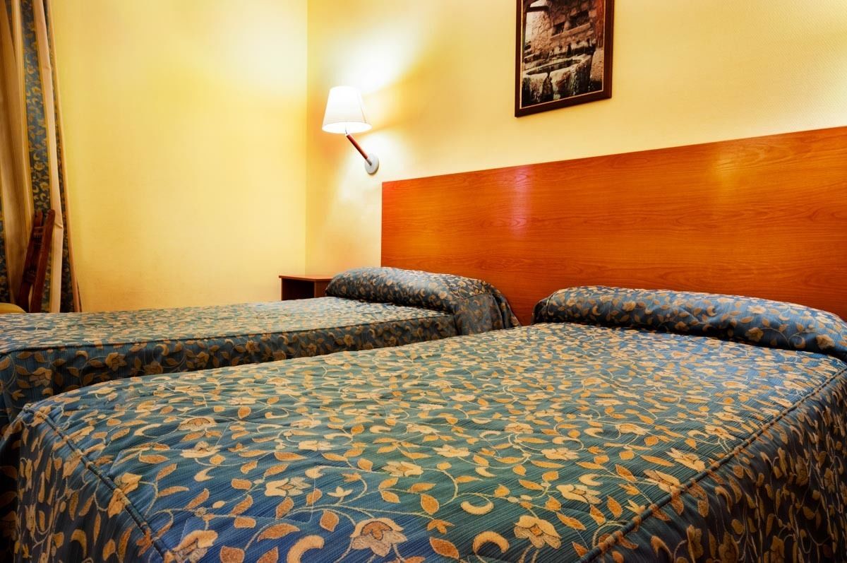 Habitación de hotel con dos camas, cabecera de madera, lámpara de pared y un cuadro enmarcado.