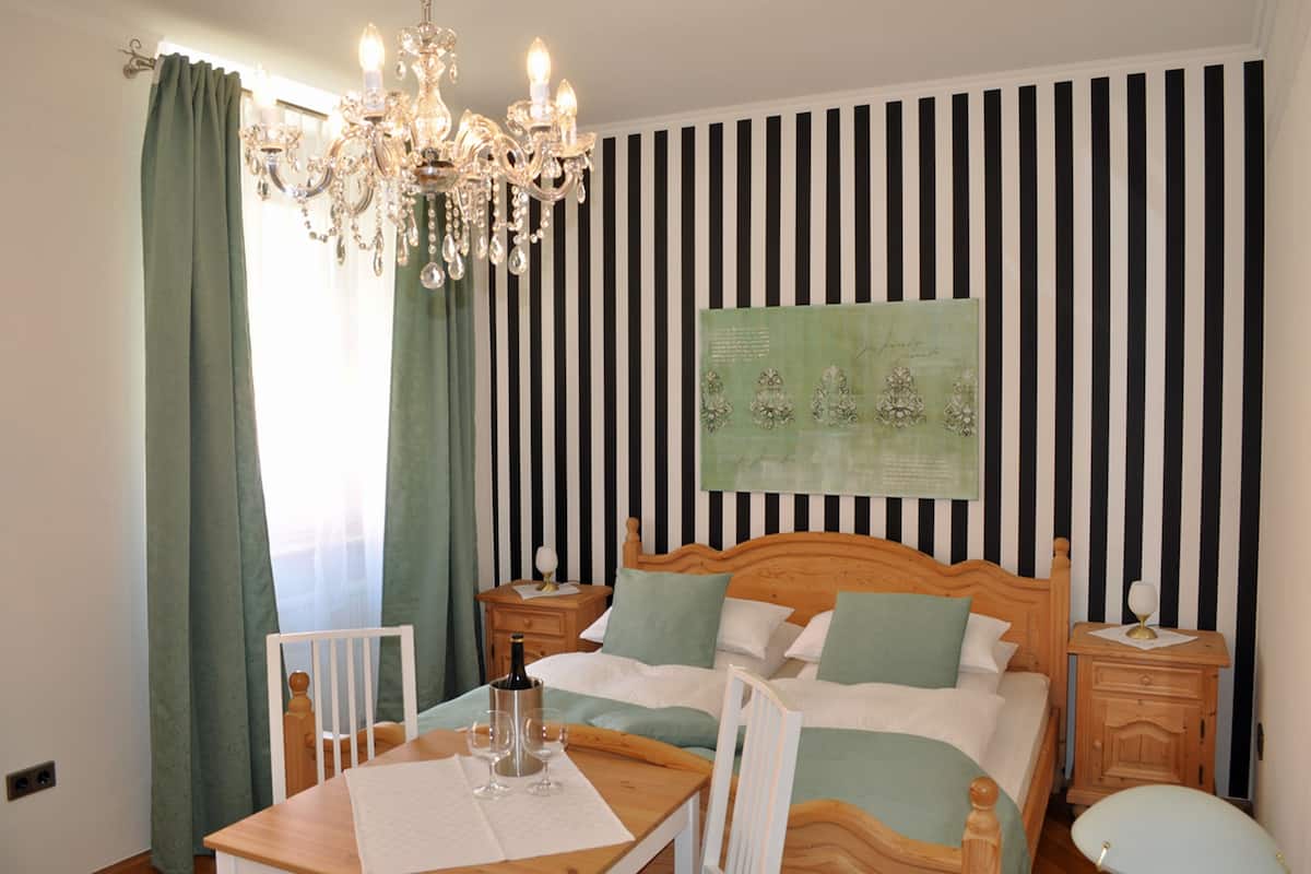A bedroom with a wooden bed, green curtains, a chandelier, a nightstand, and a table with glasses and a bottle.