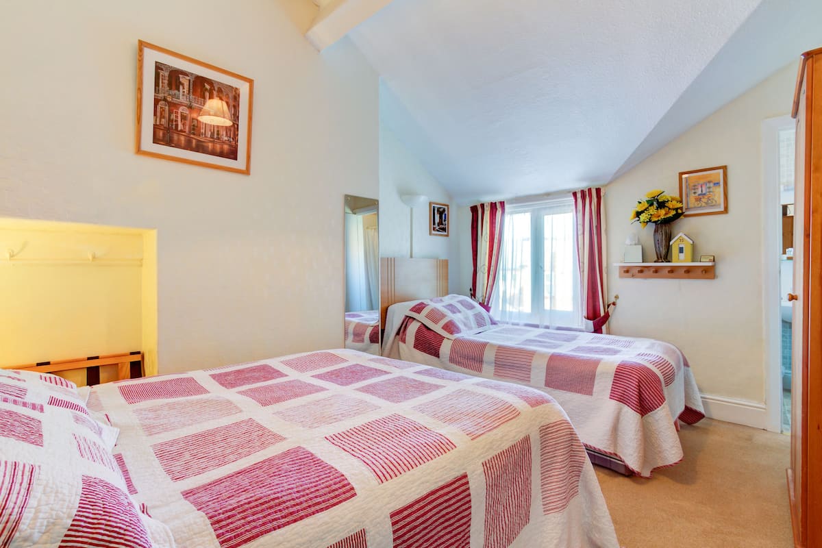 A bedroom with two beds, a window with curtains, a mirror, and a shelf with flowers.
