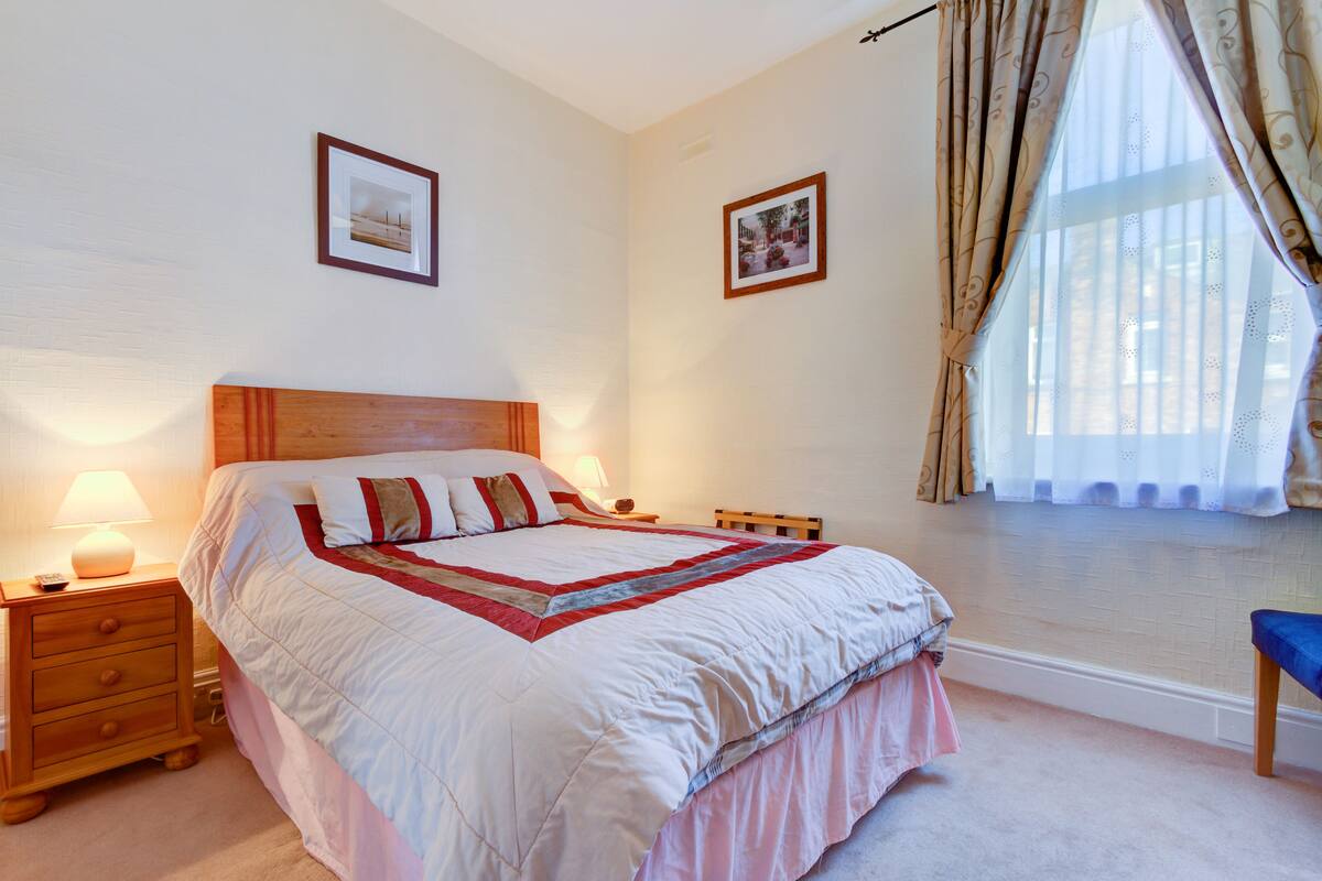 A neatly made bed with a wooden headboard, a bedside table with a lamp, and a window with curtains.