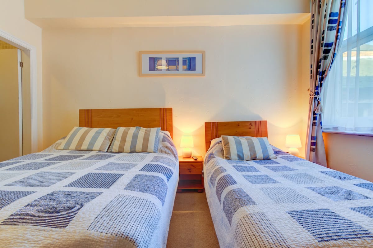 A bedroom with two beds, each with a wooden headboard and striped bedding. There is a framed picture on the wall above the beds and a window with curtains to the right.
