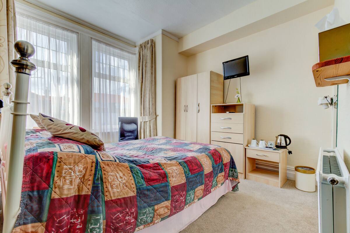 A bedroom with a large bed, a TV mounted on the wall, a desk with drawers, and a window with curtains.