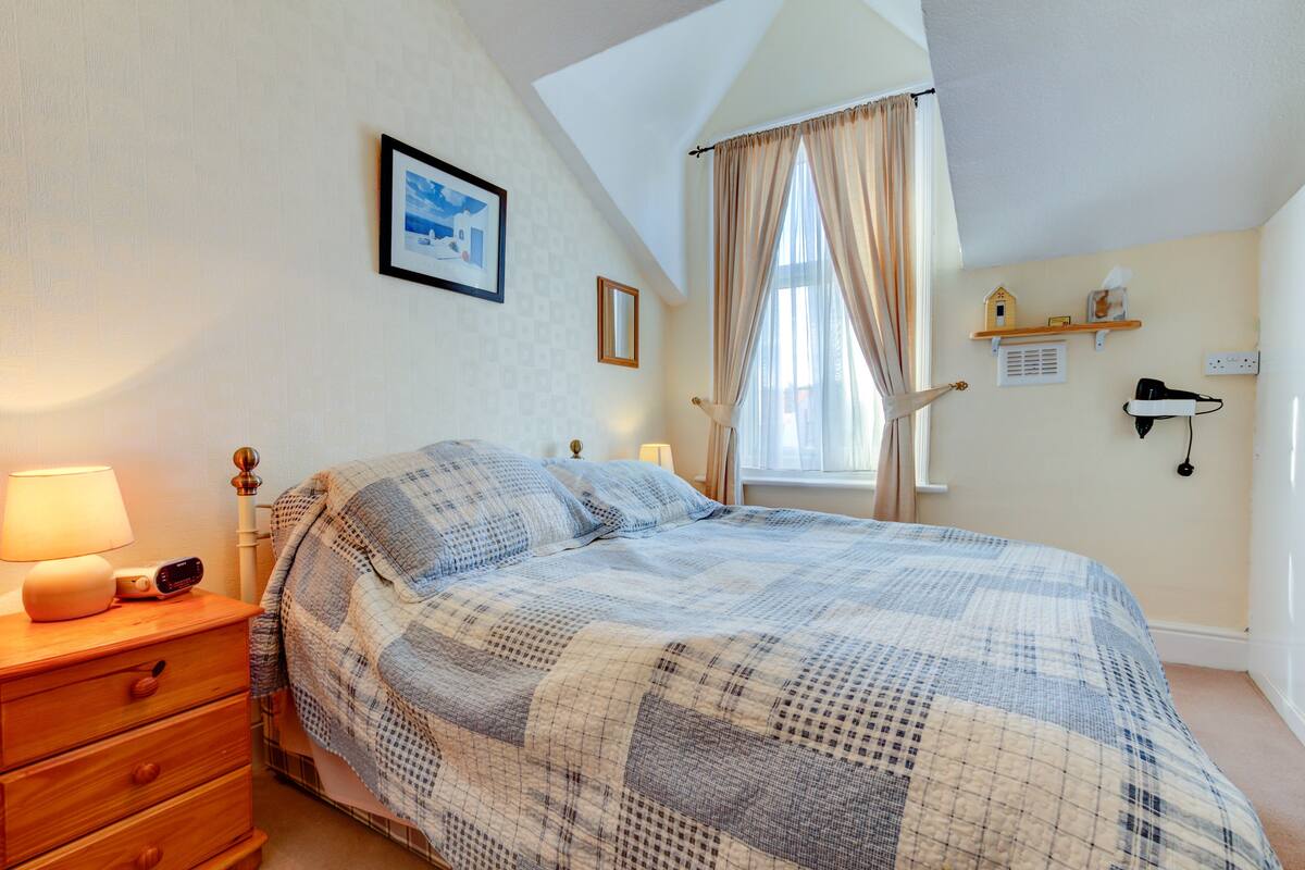 A bedroom with a bed, a nightstand, a lamp, a framed picture, a window with curtains, and a shelf with a birdhouse and a tissue box.