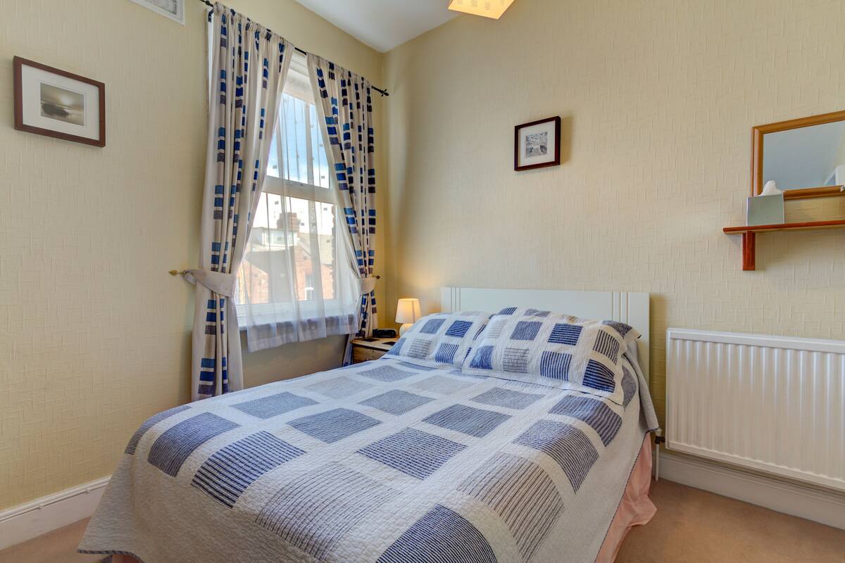 A bedroom with a bed, a nightstand, a lamp, a window with curtains, and a wall-mounted shelf.