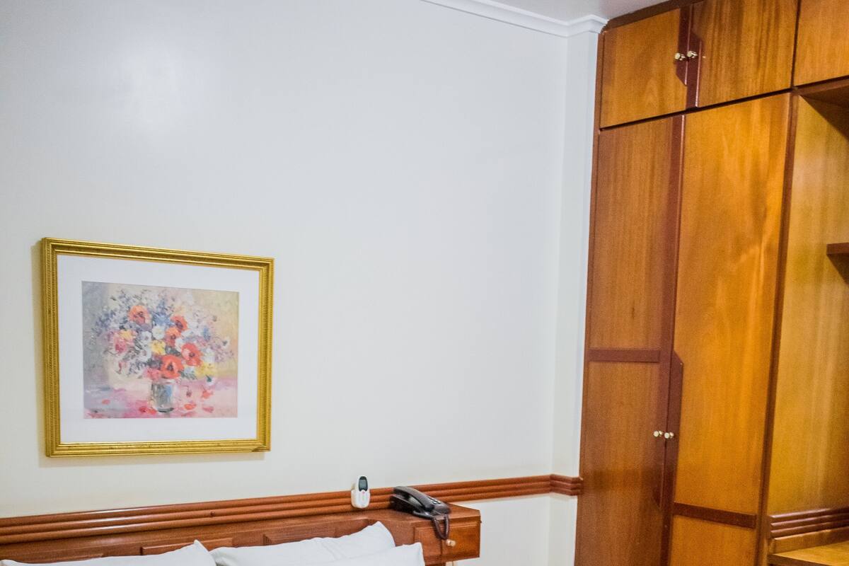 A neatly made bed with white linens and a rolled towel, a wooden wardrobe, and a framed floral painting on the wall.