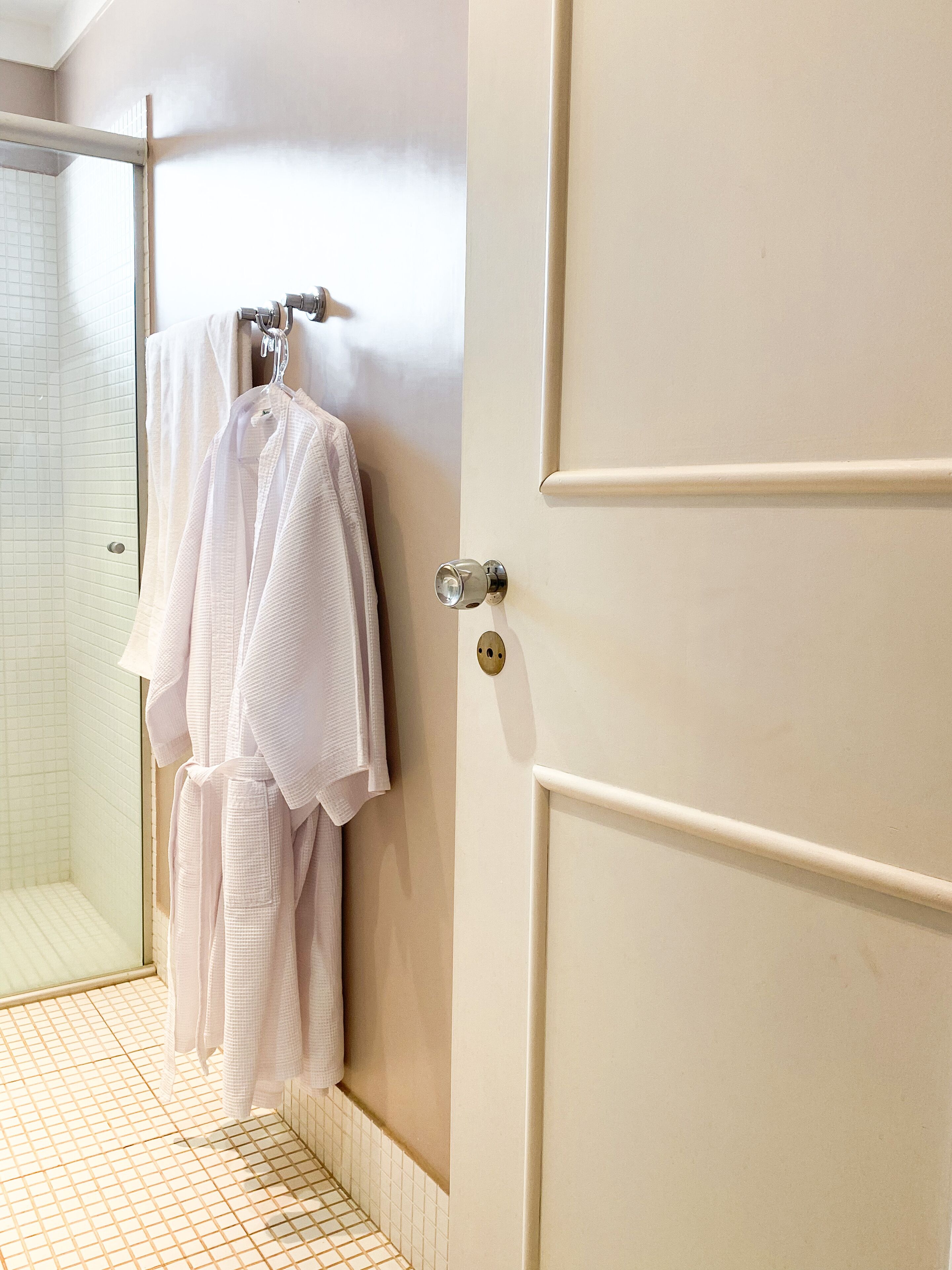 Un baño con mampara de vidrio, un perchero con varias toallas y una puerta color beige con pomo.