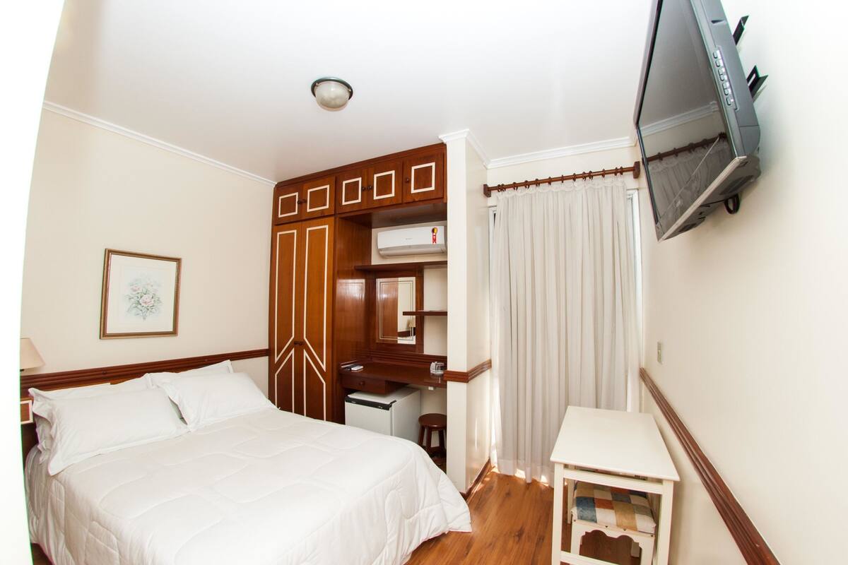A hotel room with a bed, a desk, a television mounted on the wall, and a window with curtains.