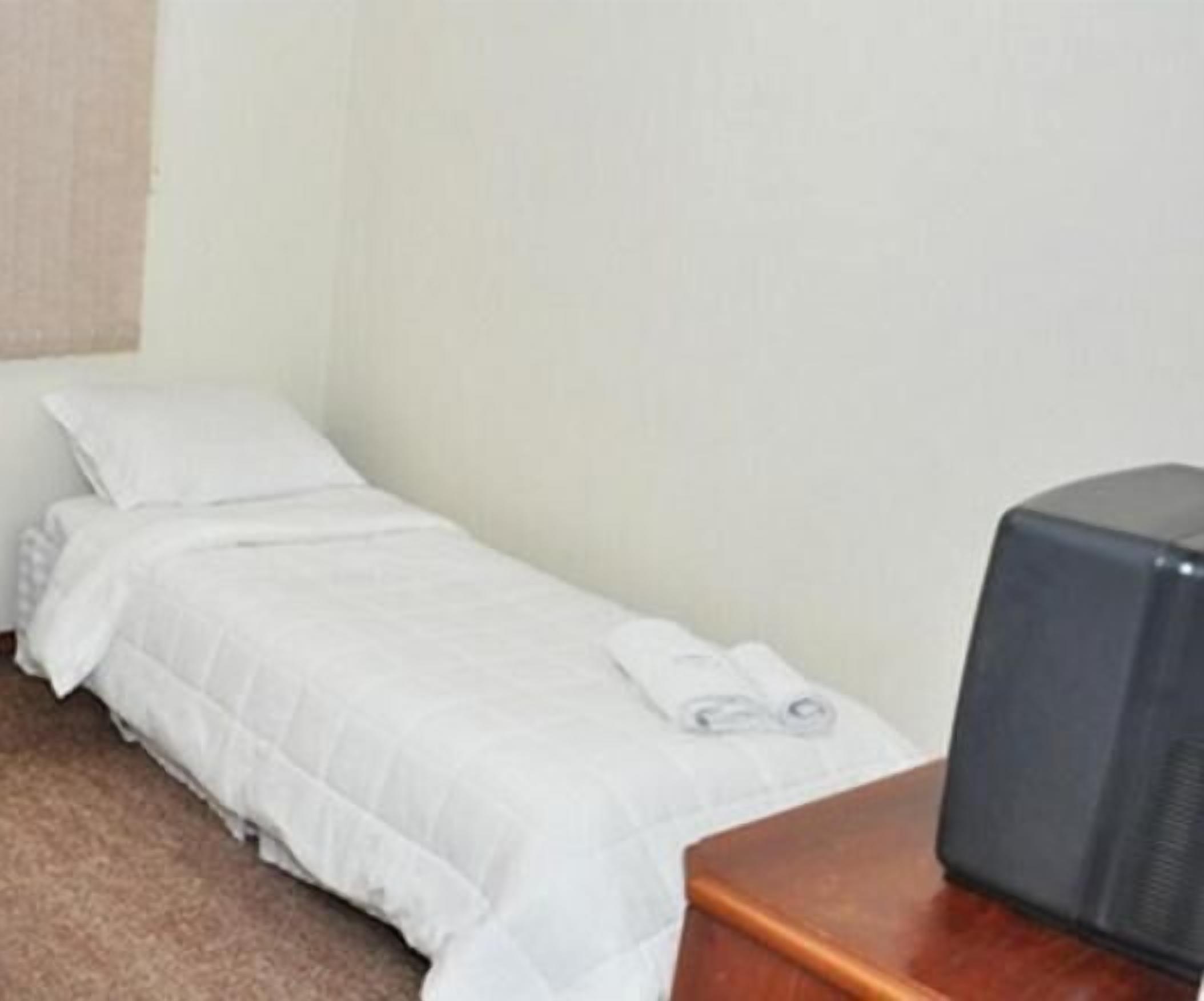 Una habitación pequeña con cama individual, una colcha blanca, una toalla enrollada y un televisor sobre una mesa de madera.