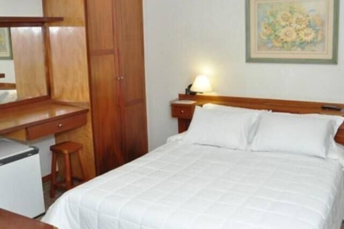 A hotel room with a bed, a desk, a chair, a wardrobe, and a nightstand with a lamp.
