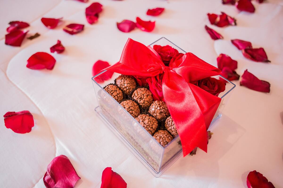 A clear glass box with a red ribbon, containing spherical chocolates, placed on a white surface with scattered rose petals.
