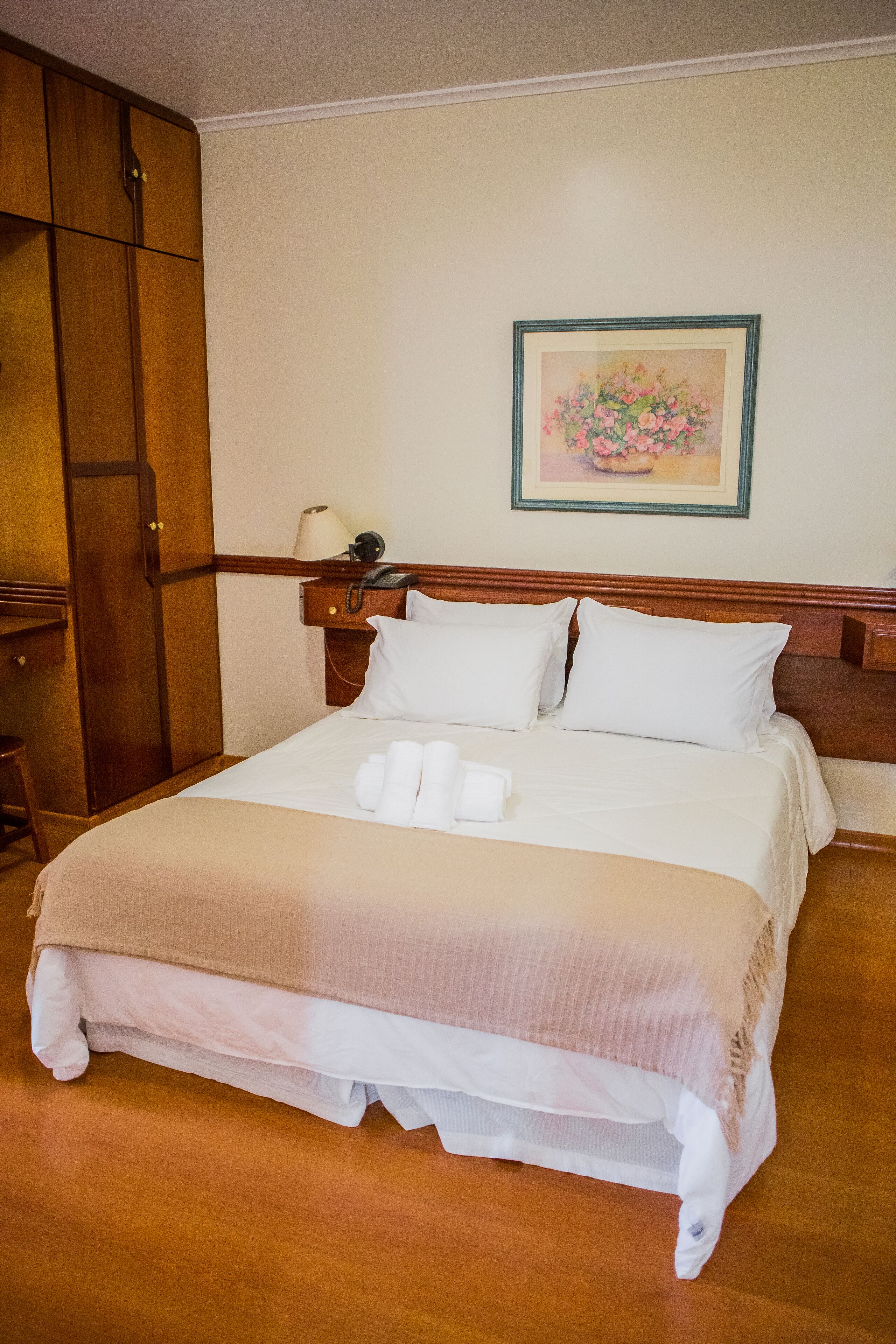 Una cama bien hecha con ropa blanca, un cabecero de madera, una mesita de noche con lámpara y un cuadro con flores en la pared.