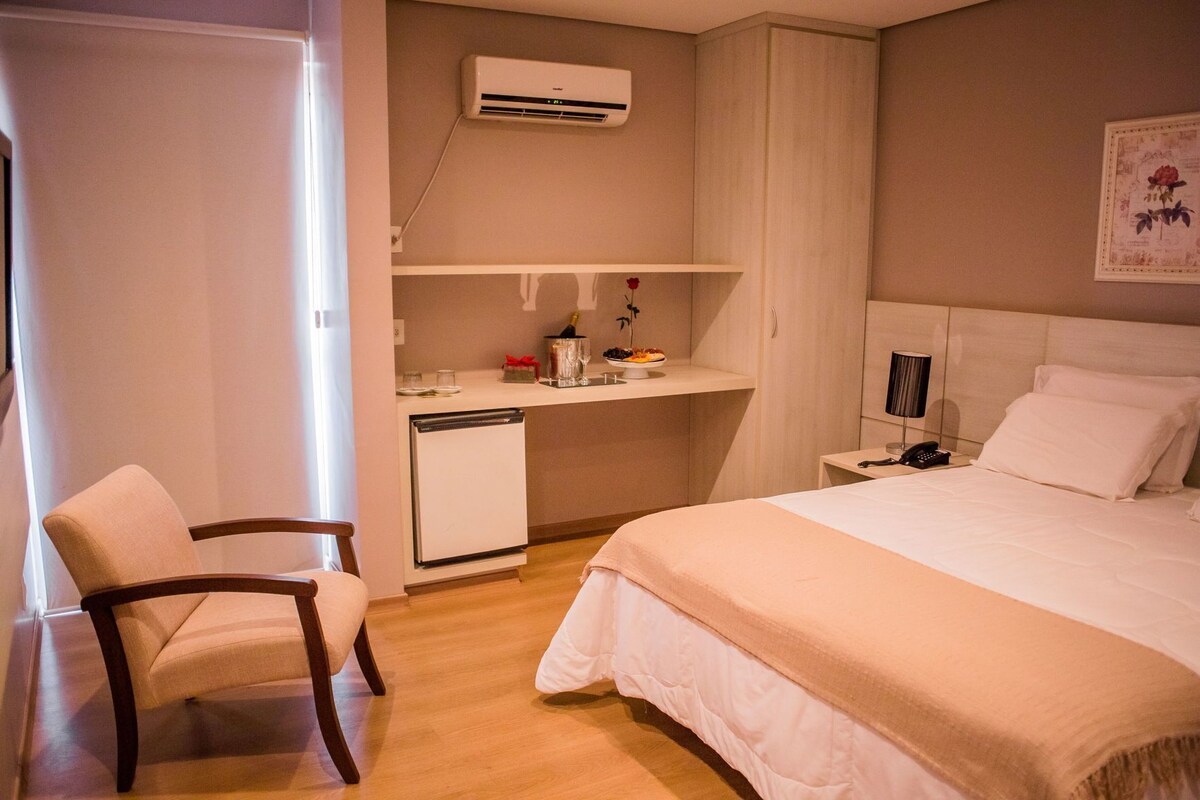 A hotel room with a bed, a desk with a chair, a small refrigerator, an air conditioner, and a bedside table with a lamp.
