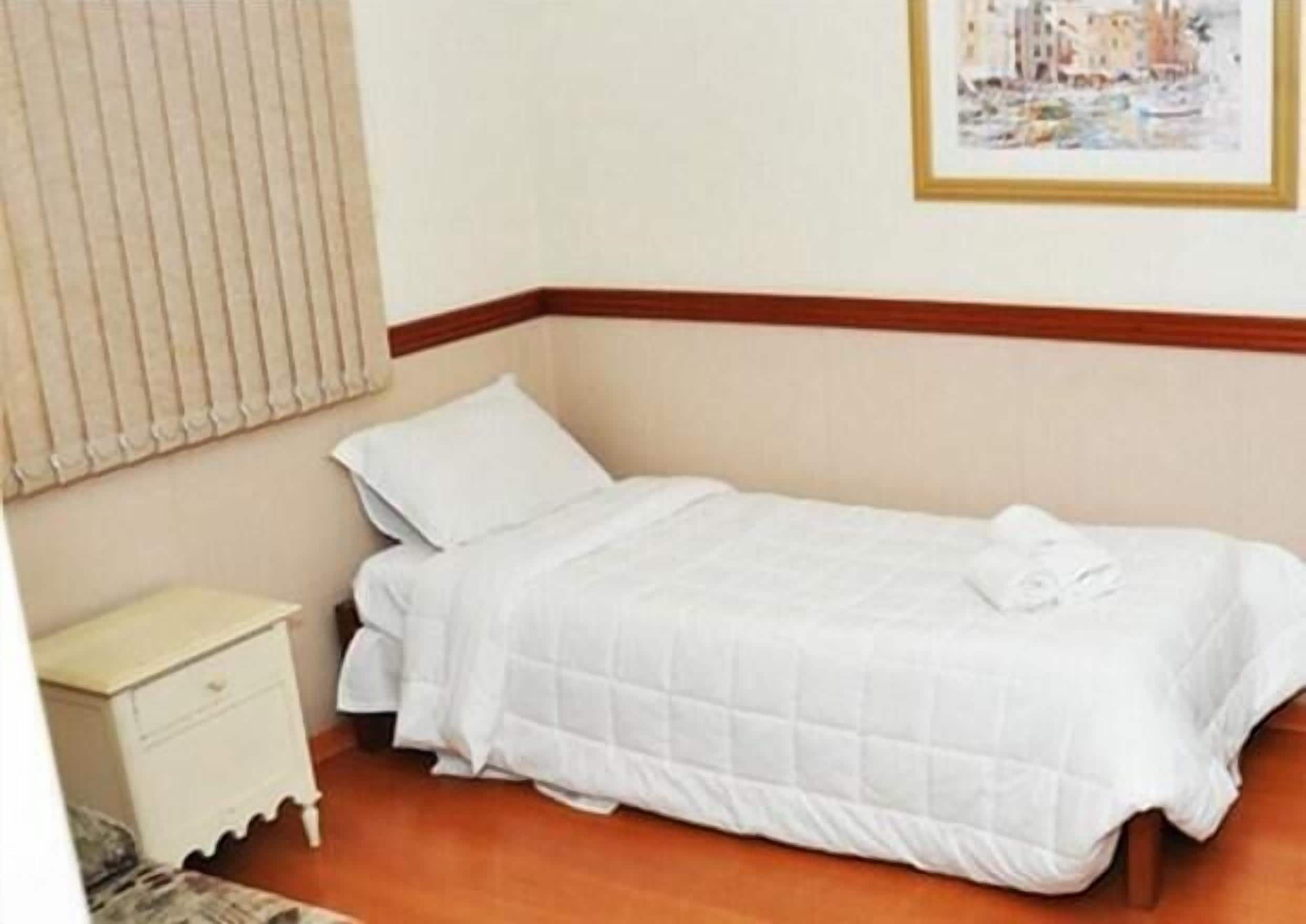 Uma cama de solteiro com colcha branca acolchoada e almofadas, cabeceira de madeira, mesa de cabeceira com gaveta e um quadro na parede.
