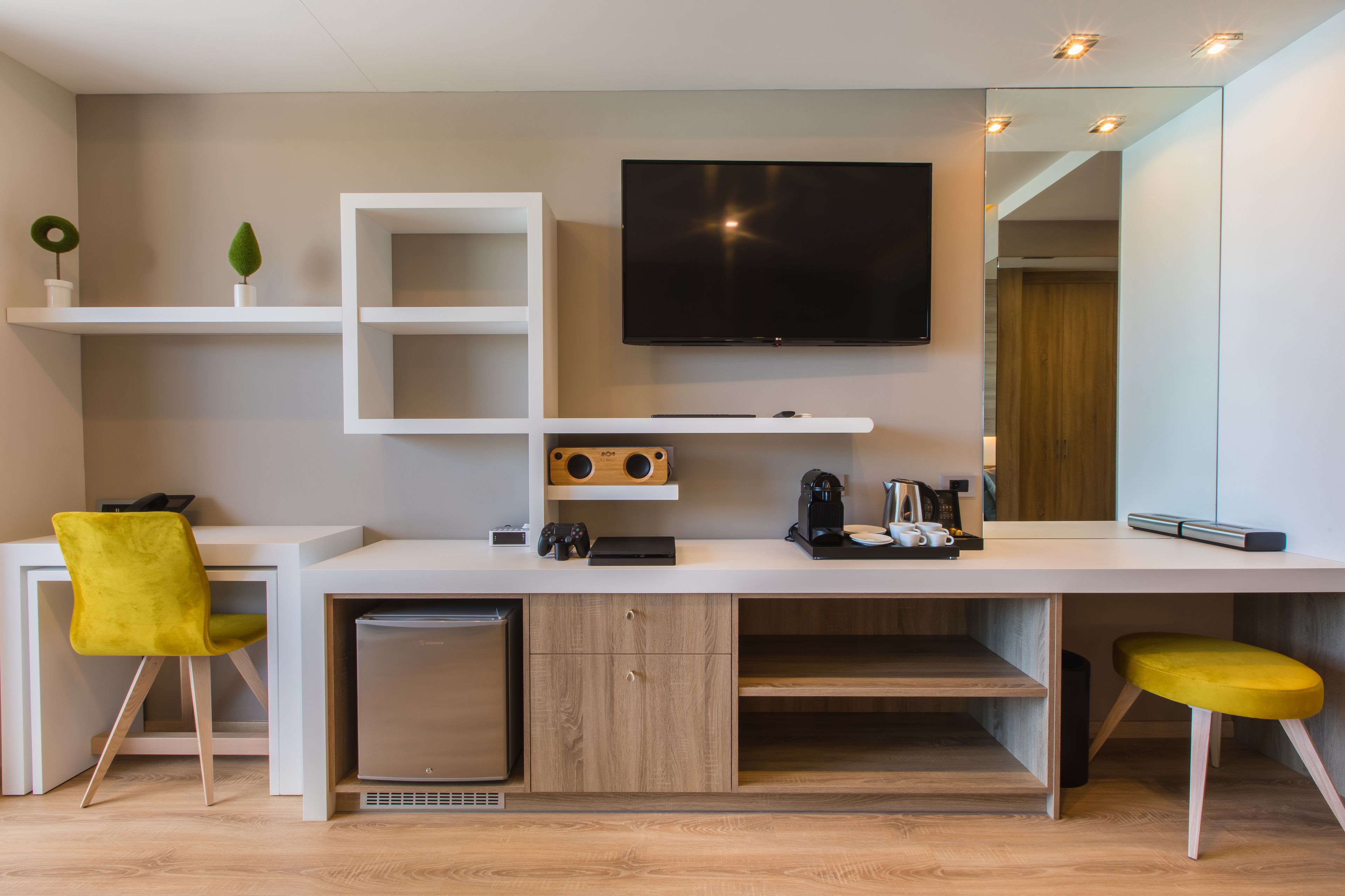 A modern room with a white desk, a flat-screen TV, a small fridge, and a yellow chair.