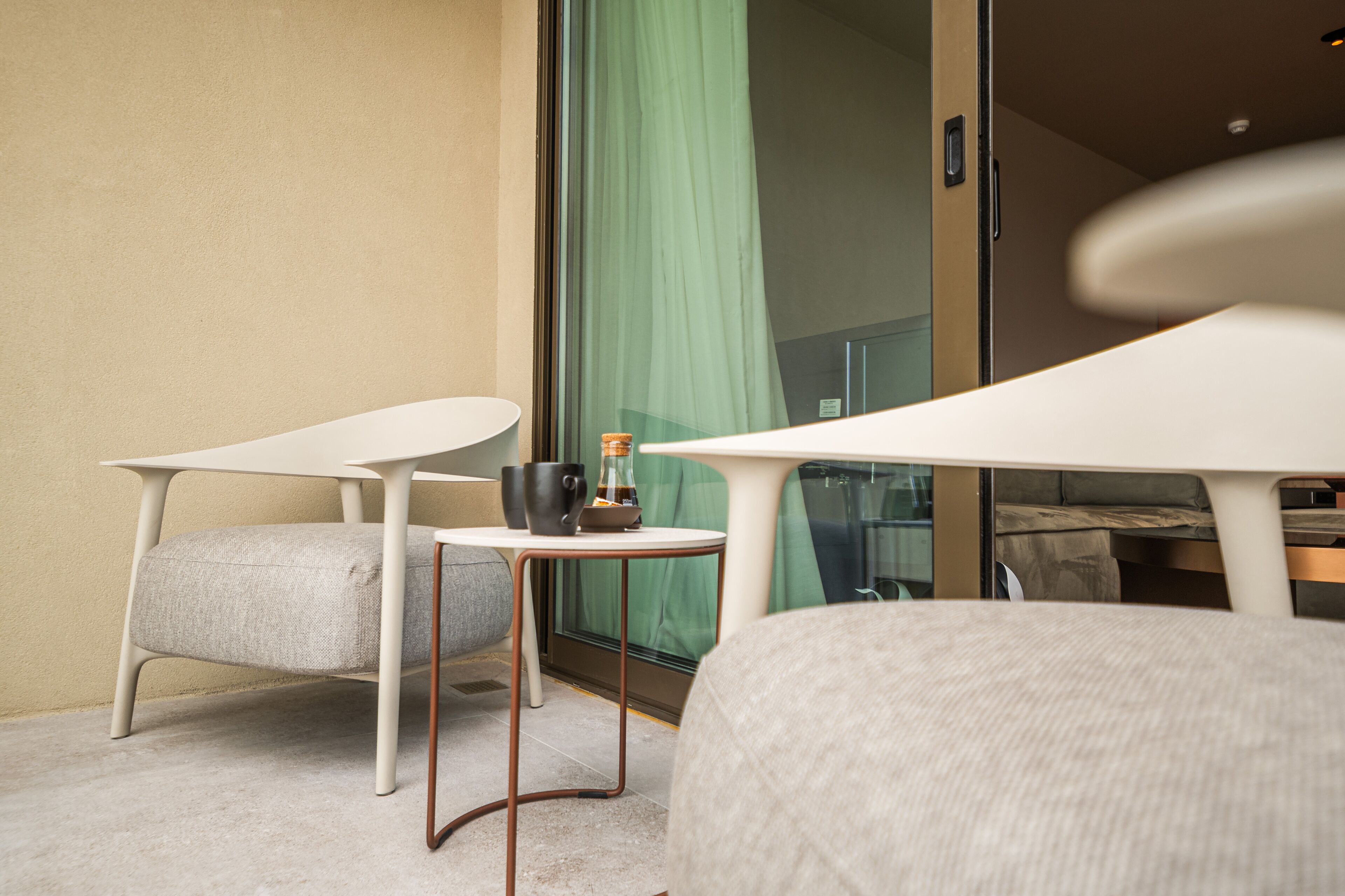 A modern hotel room with a balcony, a small table with a chair, and a view of a green curtain.