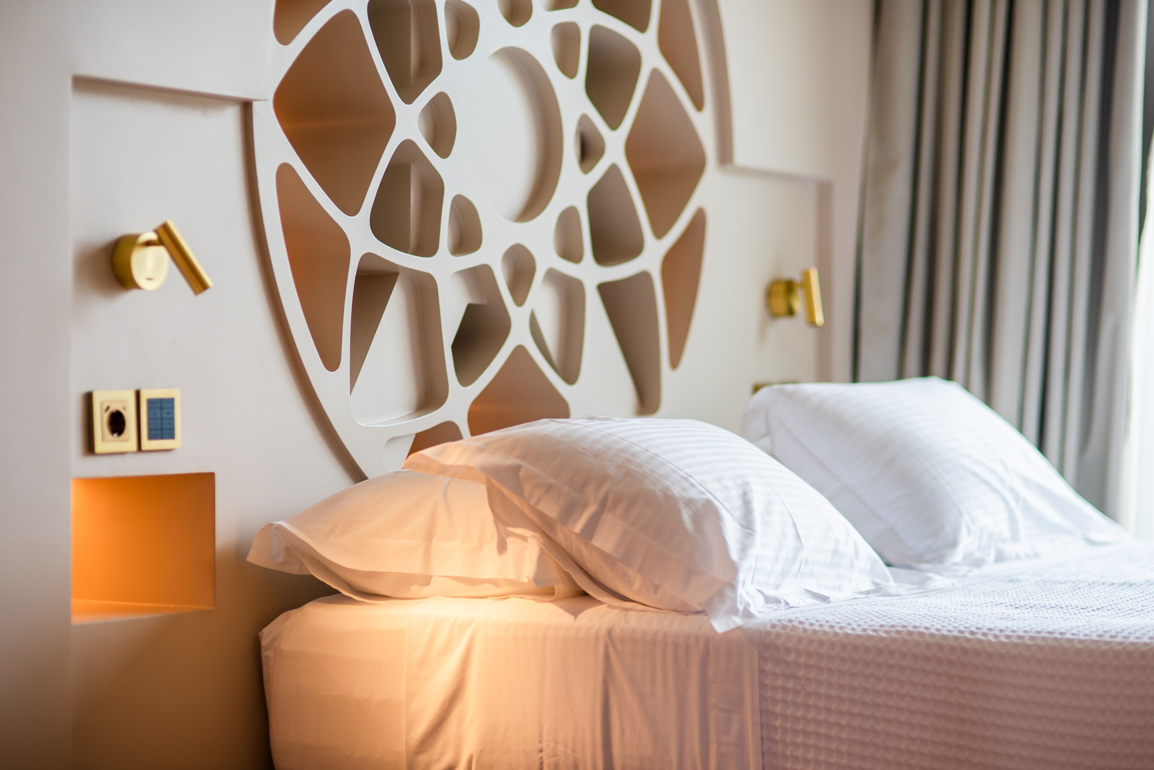 A neatly made bed with white linens and pillows, a decorative wall panel, and two wall-mounted light fixtures.