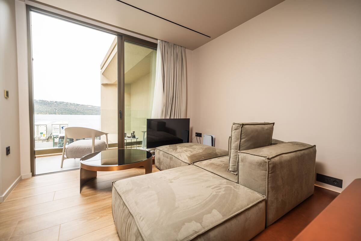 A modern living room with a sofa, a glass coffee table, and a TV. There is a sliding glass door leading to a balcony with a view of the sea.