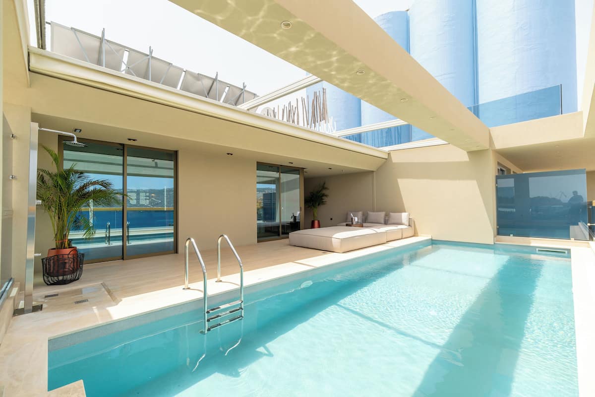 A modern indoor swimming pool with a glass balustrade, a seating area with cushions, and a view of the sea through large windows.
