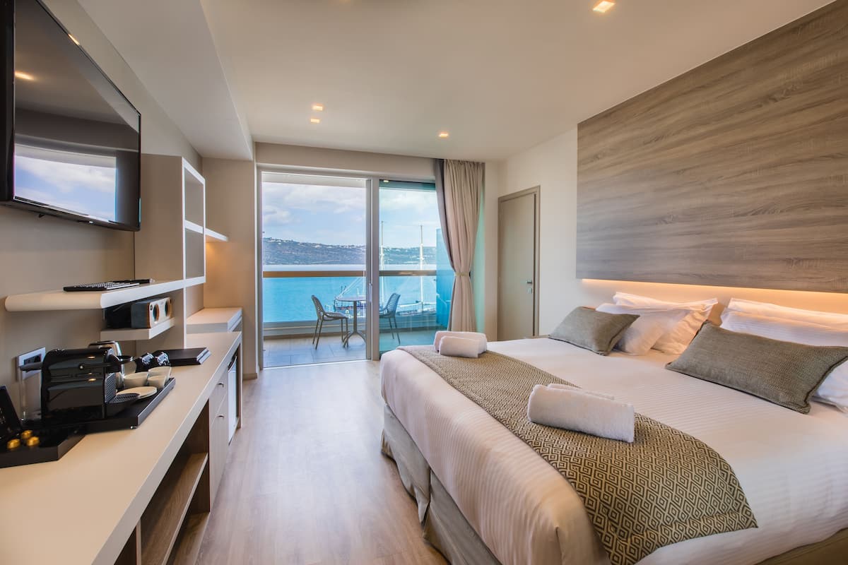 A modern hotel room with a large bed, a desk with a coffee machine, a TV, and a balcony with a view of the sea.