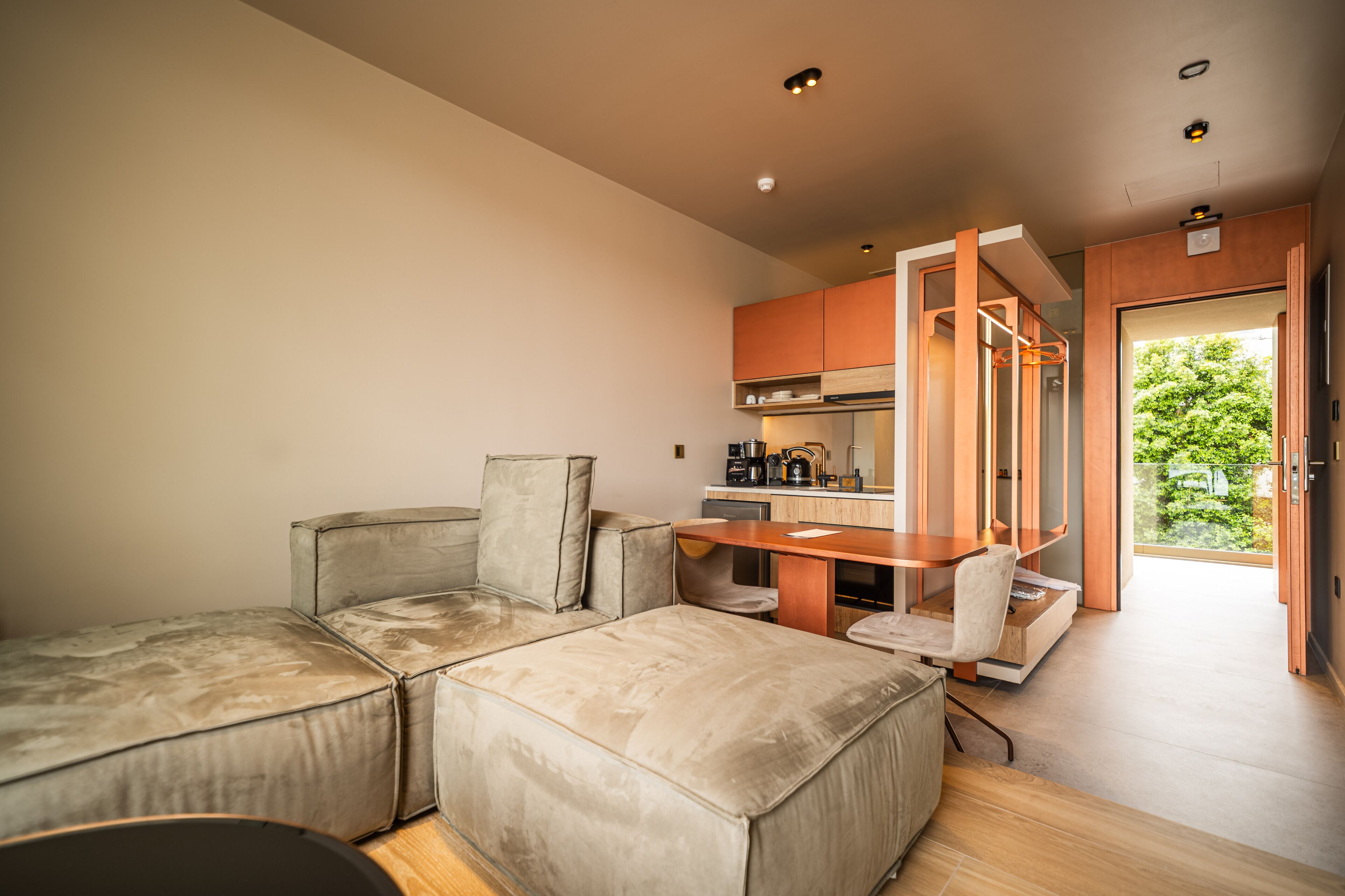 A modern living room with a sectional sofa, a small table, and a kitchen area with wooden cabinets and a coffee machine.