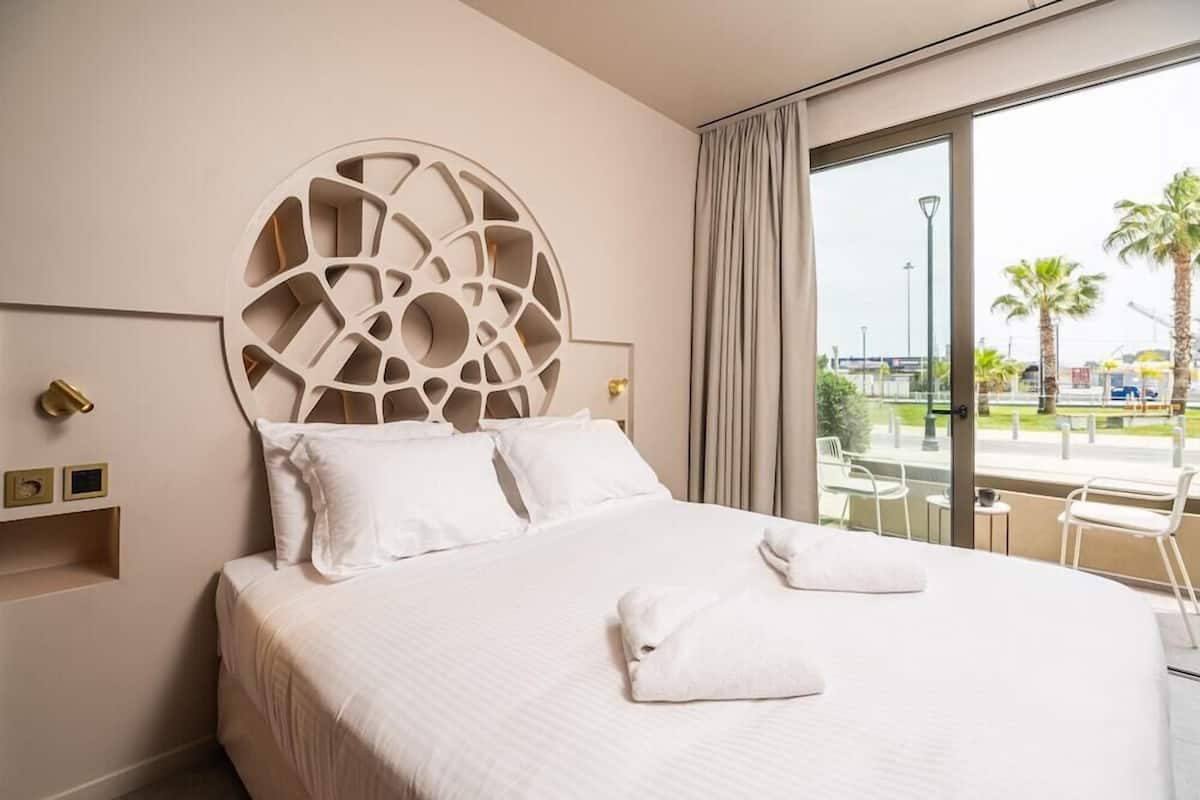 A hotel room with a large bed, a decorative headboard, a balcony with a view of palm trees and a cityscape.