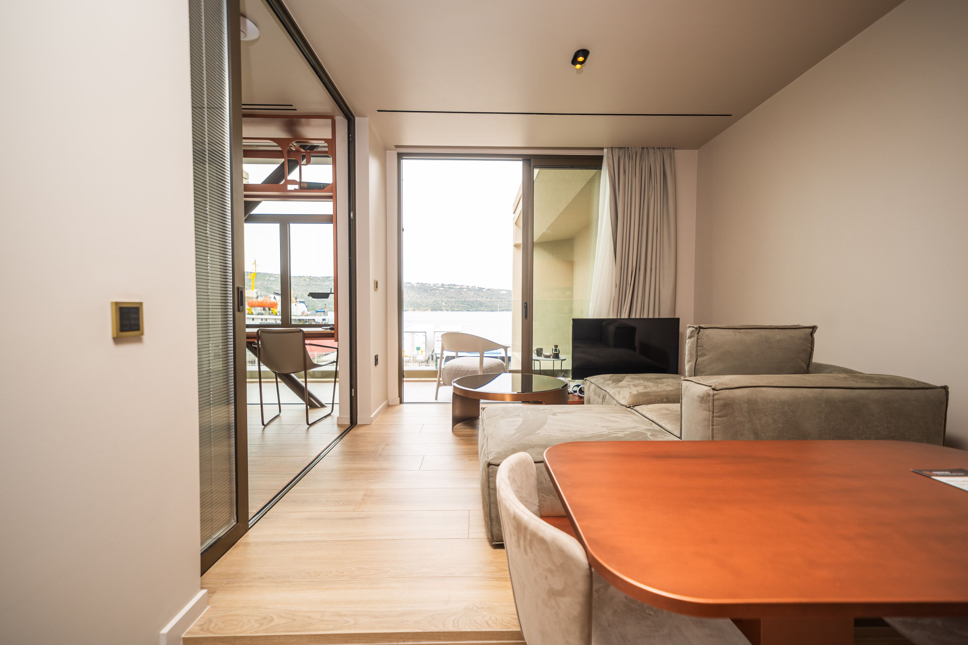 A modern living room with a sofa, a coffee table, and a TV. There is a glass door leading to a balcony with a view of the sea.