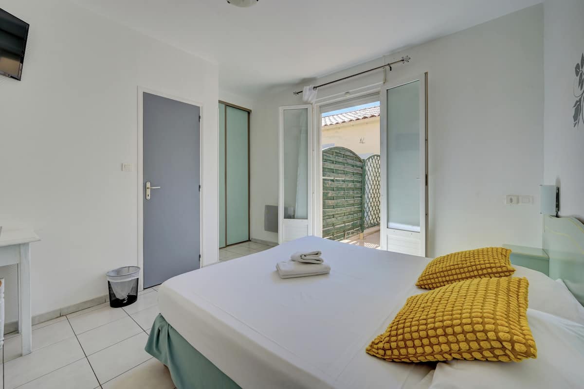 A bedroom with a bed, two pillows, a small table, a trash bin, and a sliding door leading to a balcony.