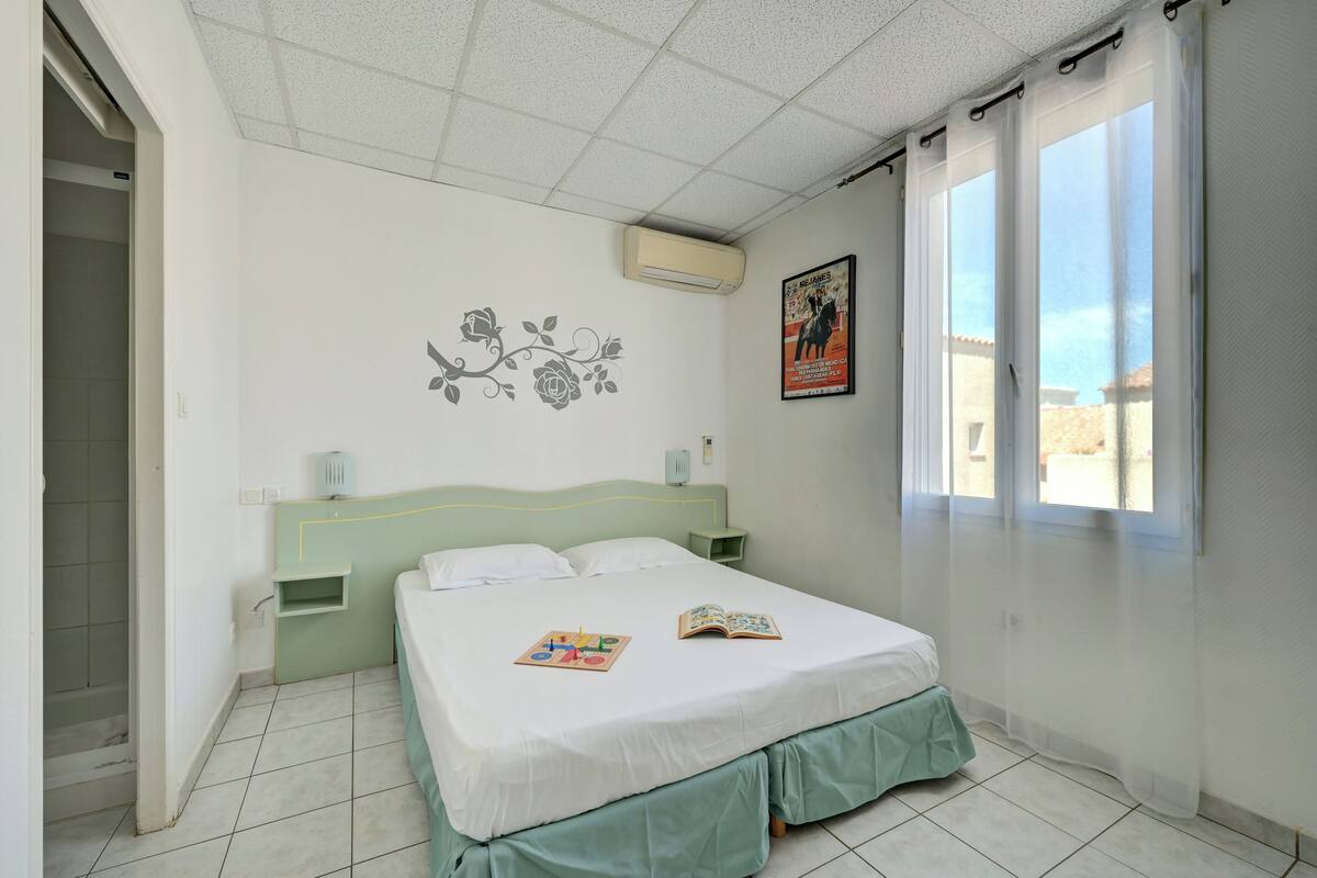 A bedroom with a bed, a nightstand, a wall-mounted air conditioner, a window with sheer curtains, and a poster on the wall.