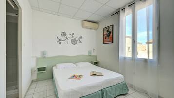 Un dormitori amb llit, una meseta de nit, aire condicionat fixat a la paret, una finestra amb cortines translúcides i un cartell a la paret.