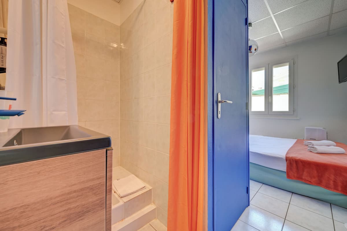 A bathroom with a shower, a sink, and a toilet. There is a bed with a red blanket and a white towel.