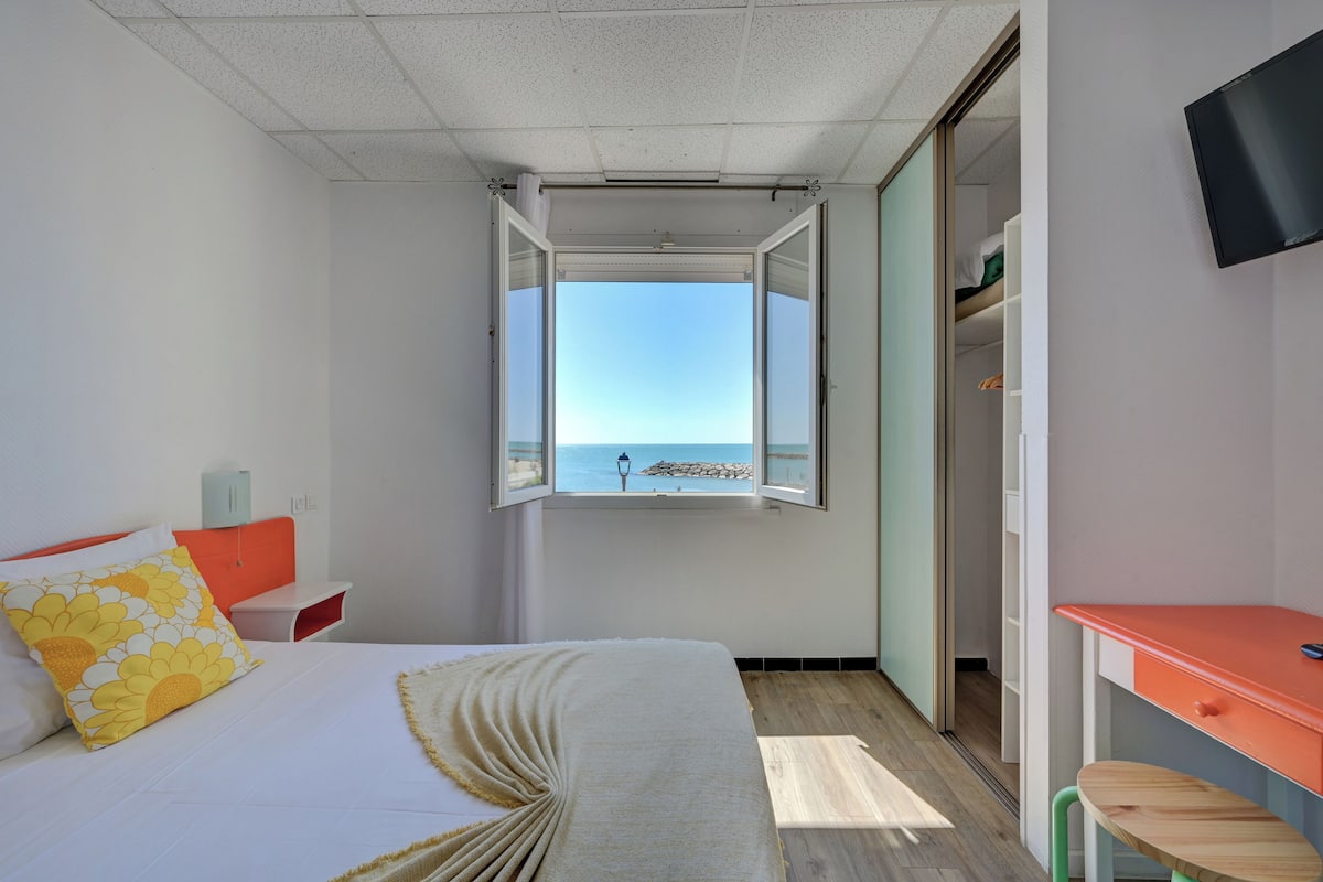 A bedroom with a bed, a desk, a chair, a wardrobe, and a window with a view of the sea.